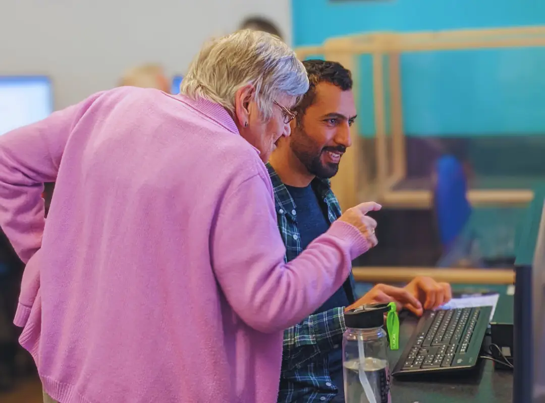 Adult male learner sits at a computer while a senior female tutor stands beside him, pointing to the screen and explaining something in a computer lab.
