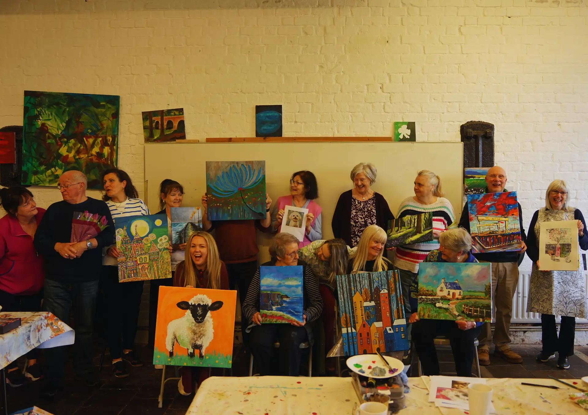 Group of fifteen adult art students standing against a white wall and holding paintings created during Conway's Intermediate Art course.