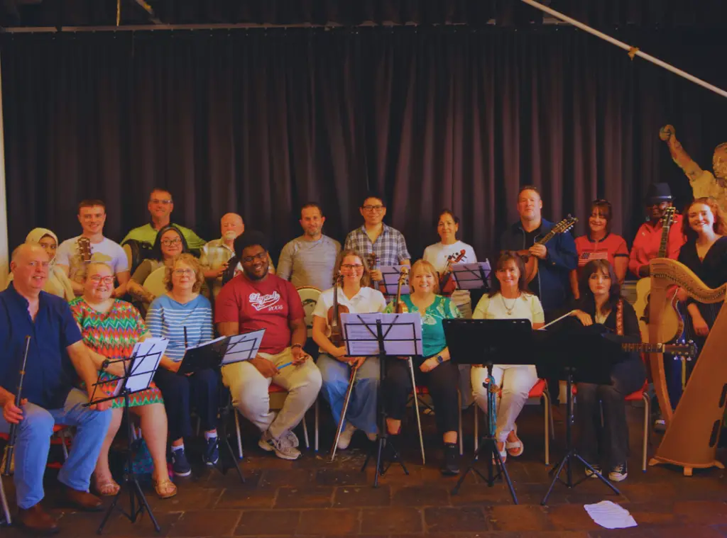 Twenty participants of the Tunes Translated music project, seated with their instruments in front of a black curtain.